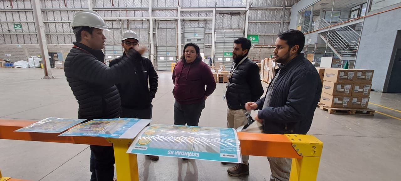 Equipo revisando en terreno láminas impresas del Estándar 5S sobre una viga amarilla, en un galpón industrial