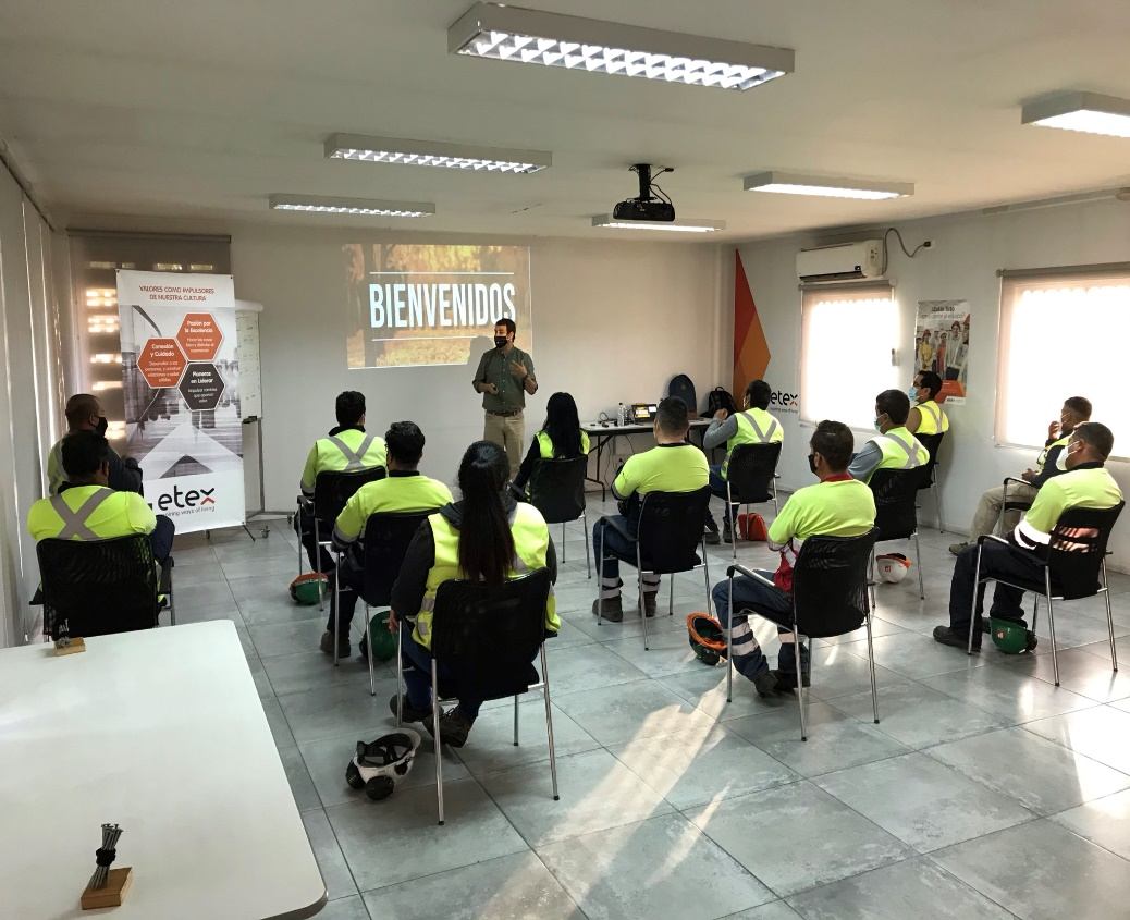 Sala de capacitación con la palabra BIENVENIDOS proyectada, equipo con chaleco reflectante sentado escuchando a un relator